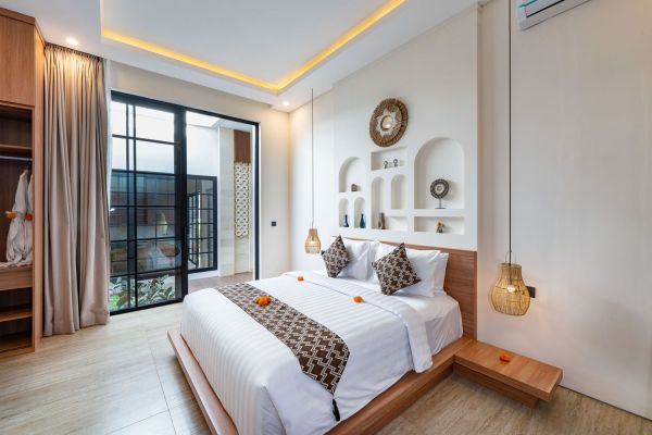 Cozy Balinese bedroom with wooden platform bed, white linens, sliding glass doors to lush garden patio, decorative wall niches, and hanging lanterns.